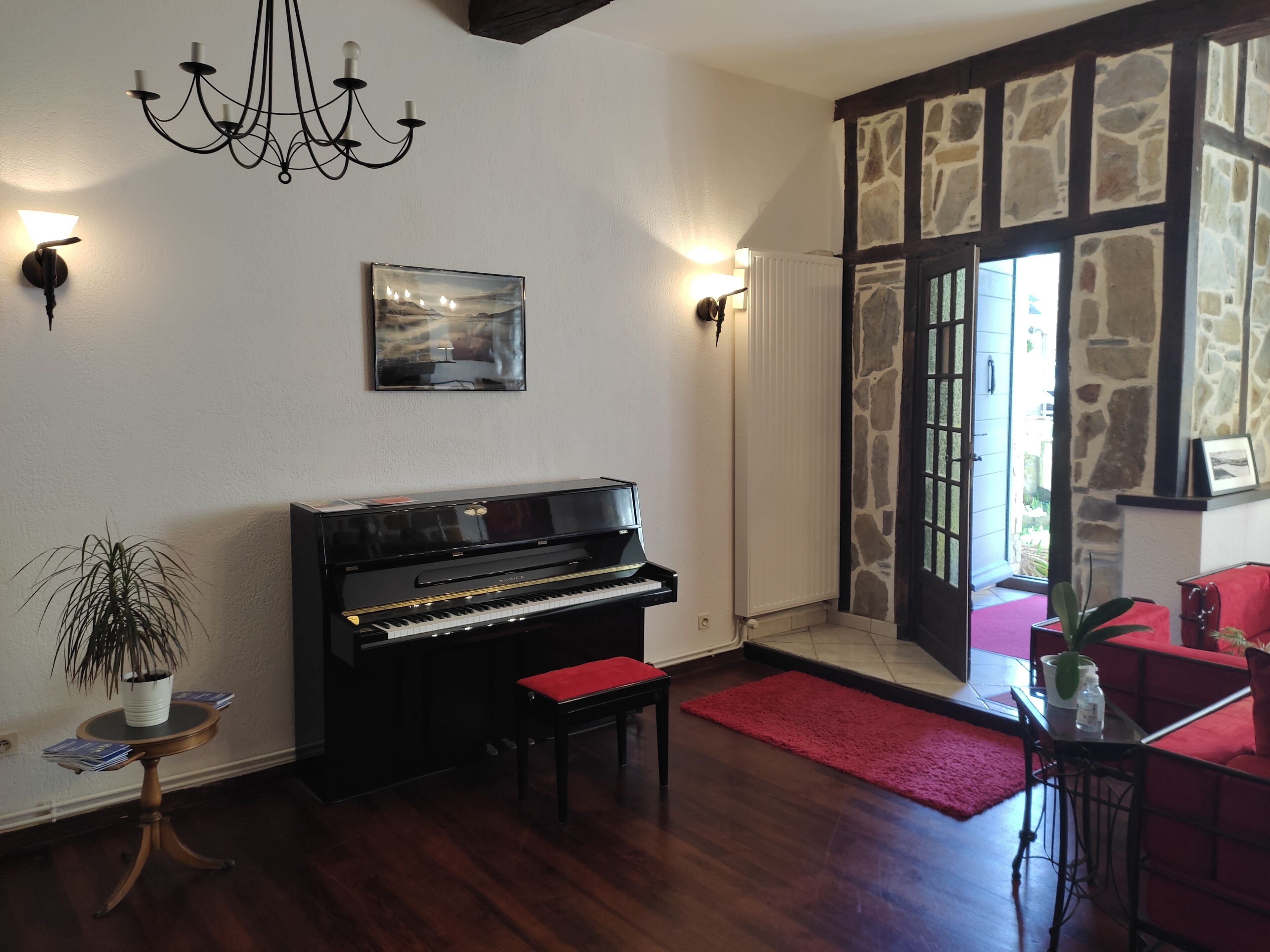 Een piano, een kroonluchter, een schilderij en een rode bank in een kamer met stenen muren en houten vloeren.