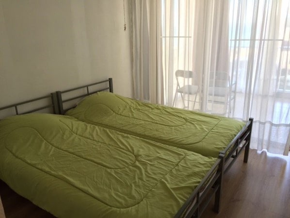 Ein Einzelbett mit grüner Bettdecke in einem Zimmer, das ein Holz-Kopfteil und ein Fenster mit durchsichtigen Vorhängen hat.