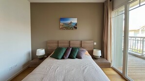 Een slaapkamer met een groot bed, een schilderij van een kustgezicht, twee bedlampjes en een uitzicht op een balkon.