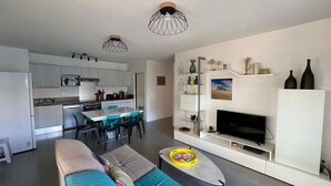 Een moderne woonkamer met een bank, salontafel en televisie. De keuken is zichtbaar, met kasten en apparatuur.