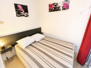 A single bed with a striped bedspread, a bedside table with a lamp, and two wall-mounted pictures. - Charming 2-room cabin with parking and pet-friendly in Sète (Sète)