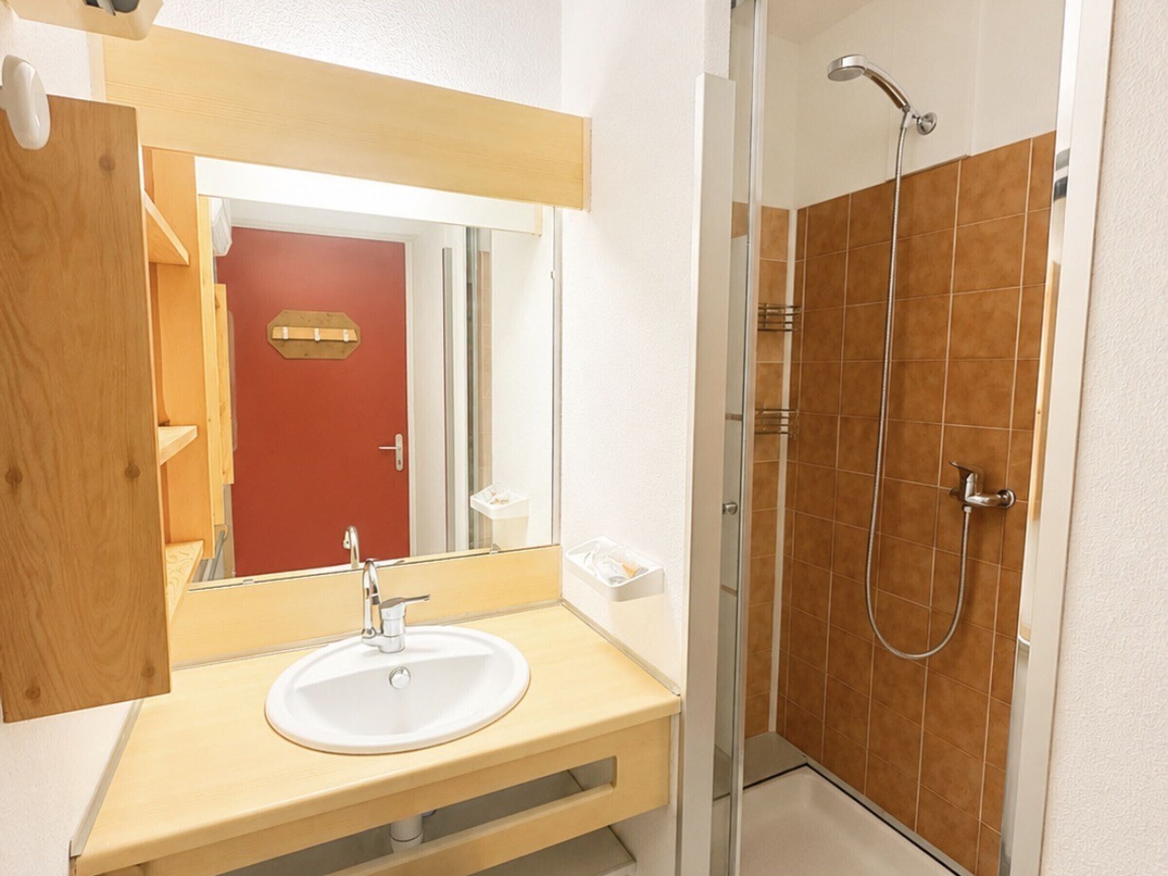 A bathroom with a sink, mirror, shower, and a red door.