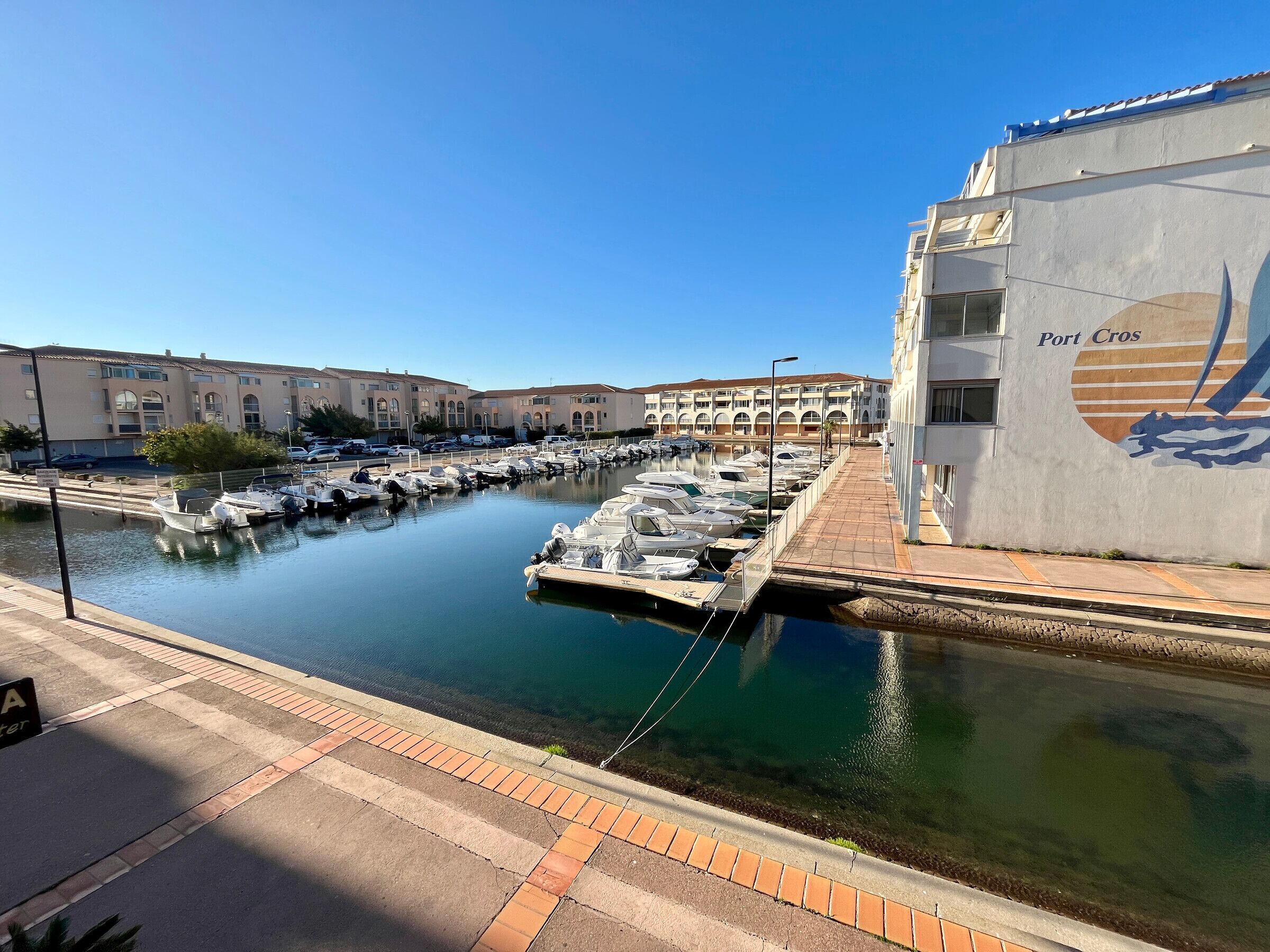 Un port de plaisance avec des bateaux amarrés, un bâtiment portant l’inscription « Port Cros » et un ciel bleu azur.