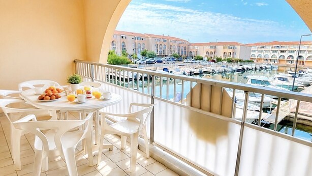 Un balcon avec une table dressée pour le petit-déjeuner, donnant sur une marina où l’on peut admirer des bateaux et des bâtiments.
