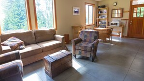 Un accogliente soggiorno con divano, poltrona e comò in legno. Sono presenti ampie finestre, una porta in legno e un angolo cottura con forno a microonde e scaffali.