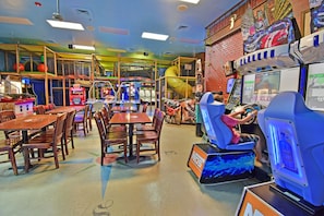 Eine Indoor-Spielehalle mit Holztischen und -stühlen, einer gelben Rutsche und verschiedenen Spielautomaten.