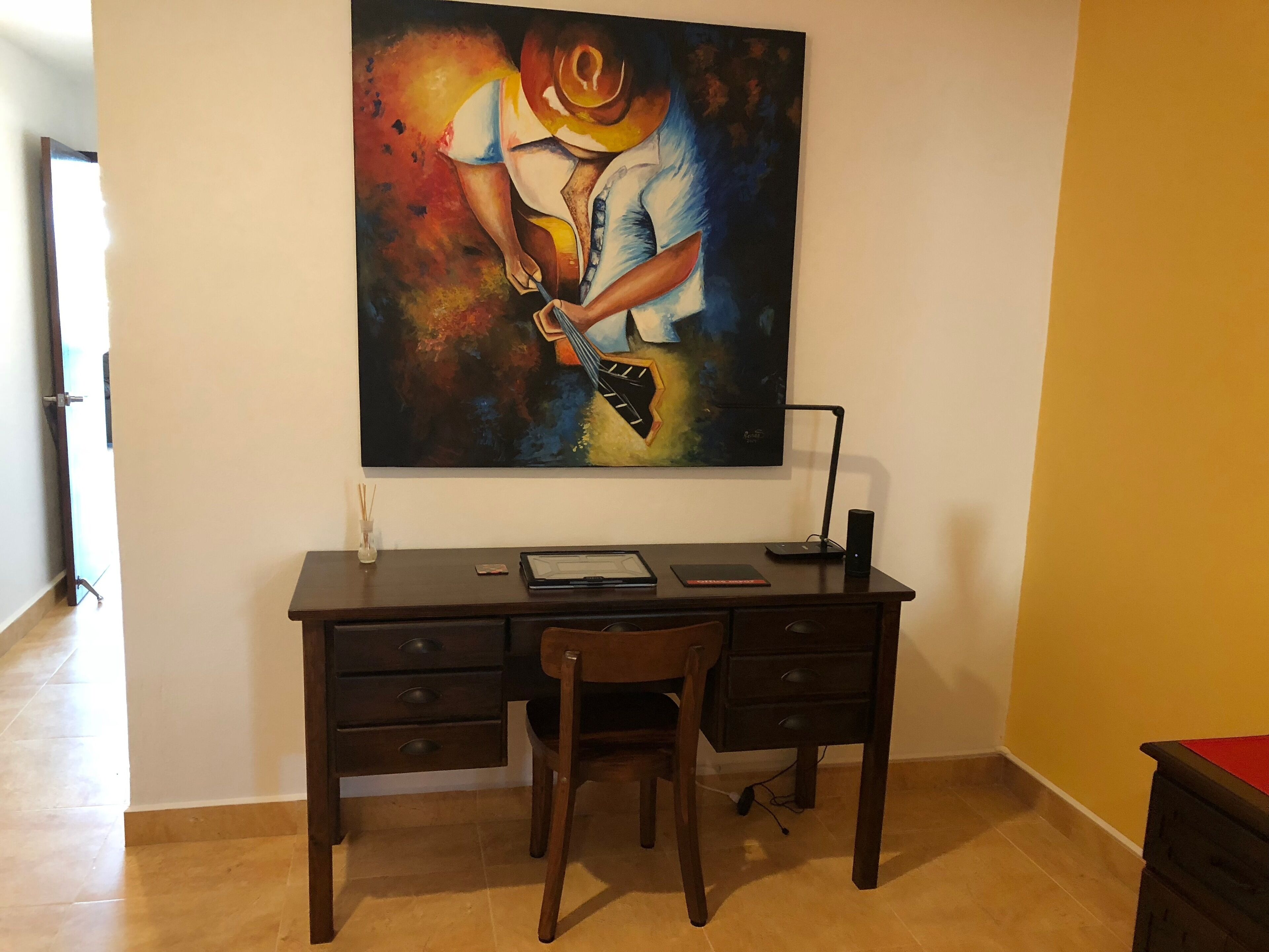 A desk with a chair, a painting on the wall, and a desk lamp.