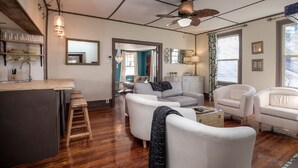 A spacious living area with a wooden floor, white sofas, a kitchen with a bar area, and a ceiling fan. - Cottage On Main- In The Heart Of Weston (Weston)