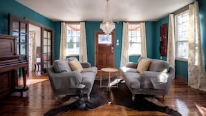 A room with teal walls, a chandelier, two grey armchairs, a wooden piano, and a cowhide rug. - Cottage On Main- In The Heart Of Weston (Weston)
