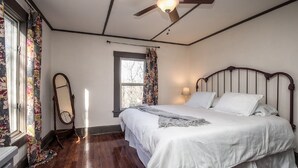 A bedroom with a wooden bed, a ceiling fan, a window with floral curtains, and a bedside lamp. - Cottage On Main- In The Heart Of Weston (Weston)