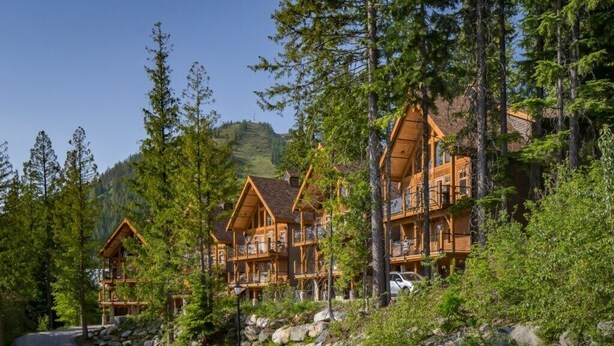 A multi-story log cabin with a stone foundation, surrounded by dense forest and a mountain backdrop. - Ski In, Ski Out Awesomeness! (Rossland)