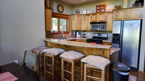 A kitchen with wooden cabinets, a stainless steel refrigerator, a microwave, and a stove. There are three stools with fabric seats and a trash can. - Ski In, Ski Out Awesomeness! (Rossland)