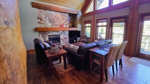 A cozy living room with a stone fireplace, wooden ceiling, and large windows. - Ski In, Ski Out Awesomeness! (Rossland)