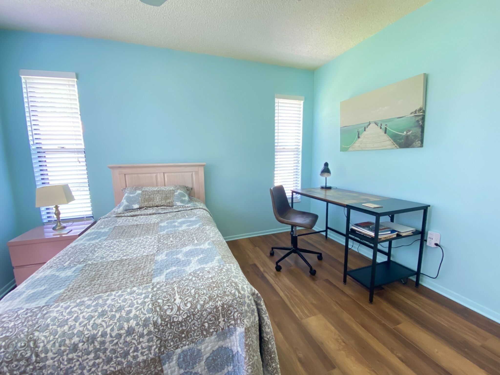 Une chambre à coucher avec un lit, un bureau et une chaise. Il y a deux fenêtres avec des stores, une lampe sur la table de chevet et un tableau au mur.