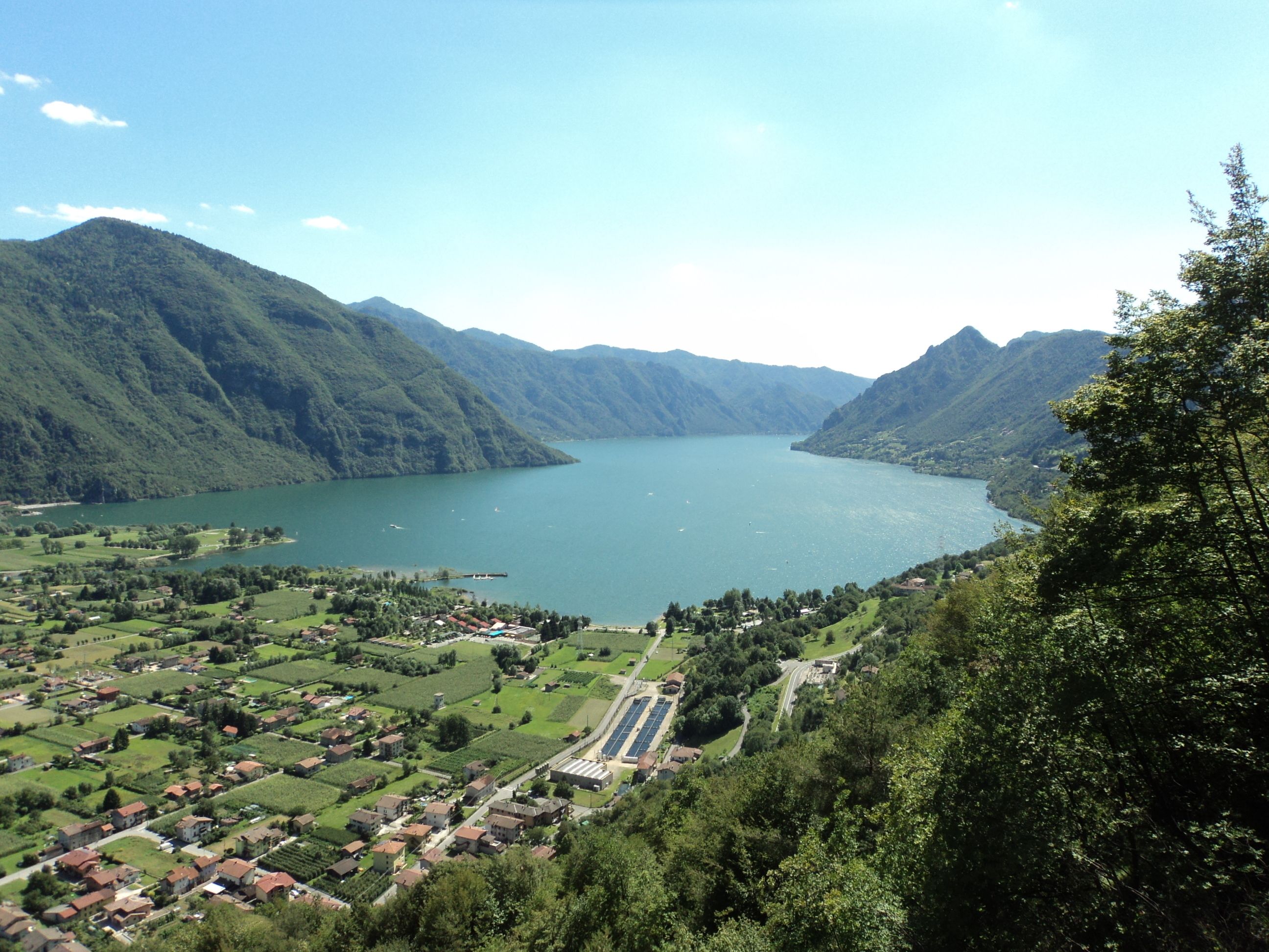 Una vista panorámica de un lago rodeado de exuberantes colinas verdes y un pequeño pueblo con casas tradicionales y una planta de energía solar.