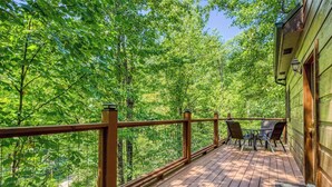 A wooden deck with a table and chairs, surrounded by a metal railing, overlooking a dense forest. - Private Wooded Setting! Theater Room! Centrally Located! 3 Decks! Fun Game Room  (Sevierville)