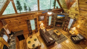 A cozy cabin interior with a dining area, sofa, fireplace, and large windows overlooking a forest. - Private Wooded Setting! Theater Room! Centrally Located! 3 Decks! Fun Game Room  (Sevierville)