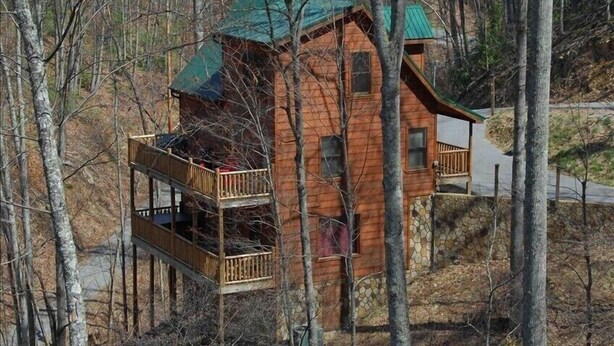 A cabin with a green roof, wooden balconies, and a stone foundation. - Private Wooded Setting! Theater Room! Centrally Located! 3 Decks! Fun Game Room  (Sevierville)