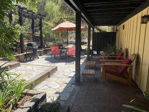 Ein überdachter Patio-Bereich mit Steinboden, Gartenmöbeln und einem Grill.