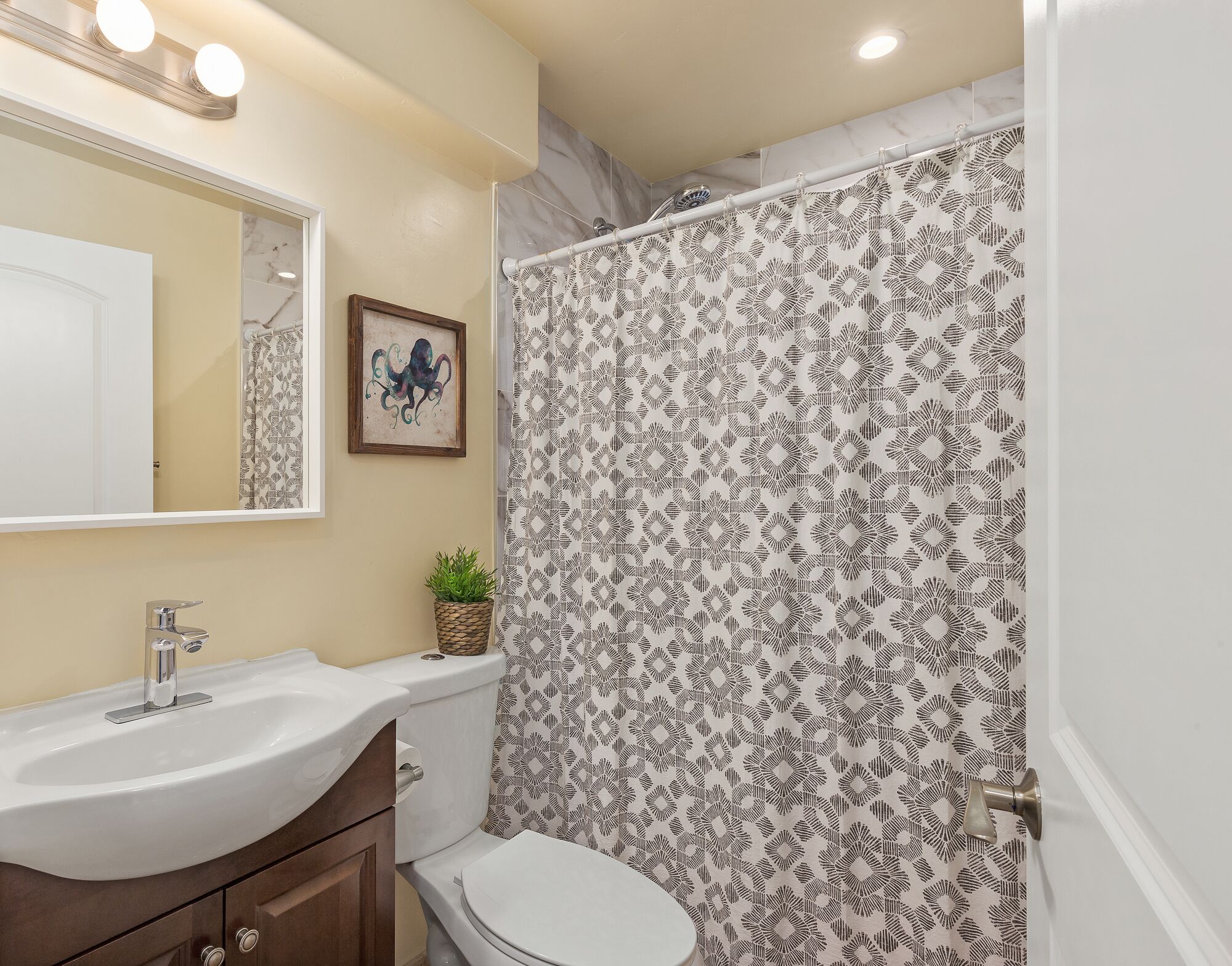Un baño con un lavamanos blanco, una obra de arte enmarco, una planta en maceta y una ducha con cortina estampada.