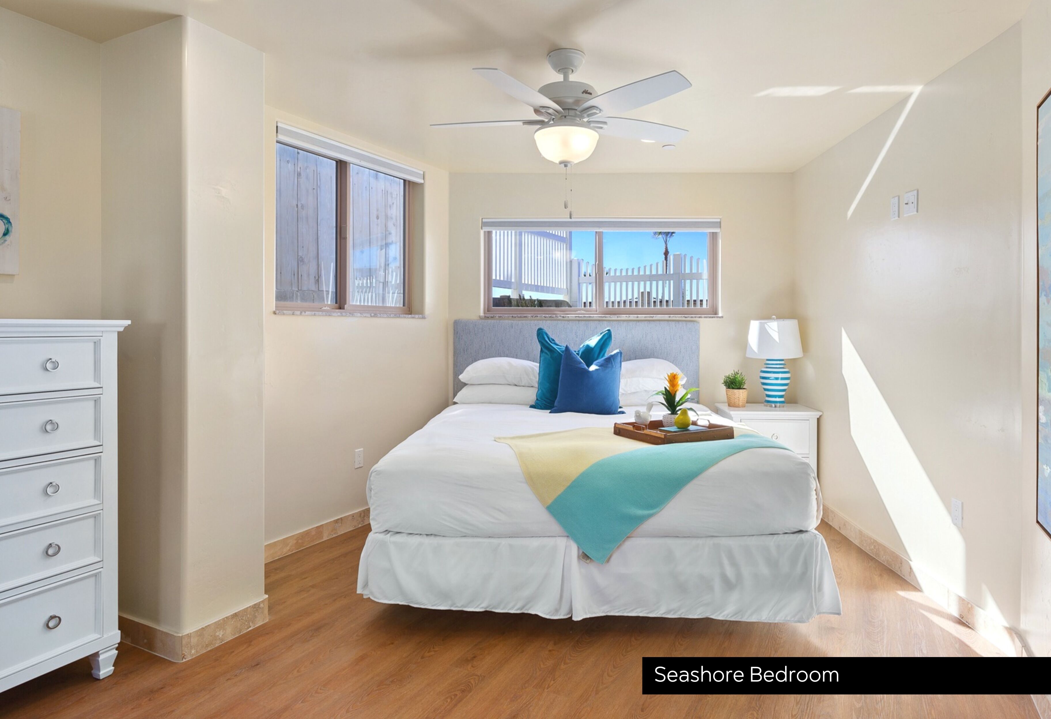 A bedroom with a bed, a dresser, a nightstand, a ceiling fan, and a window with a view of a white fence.