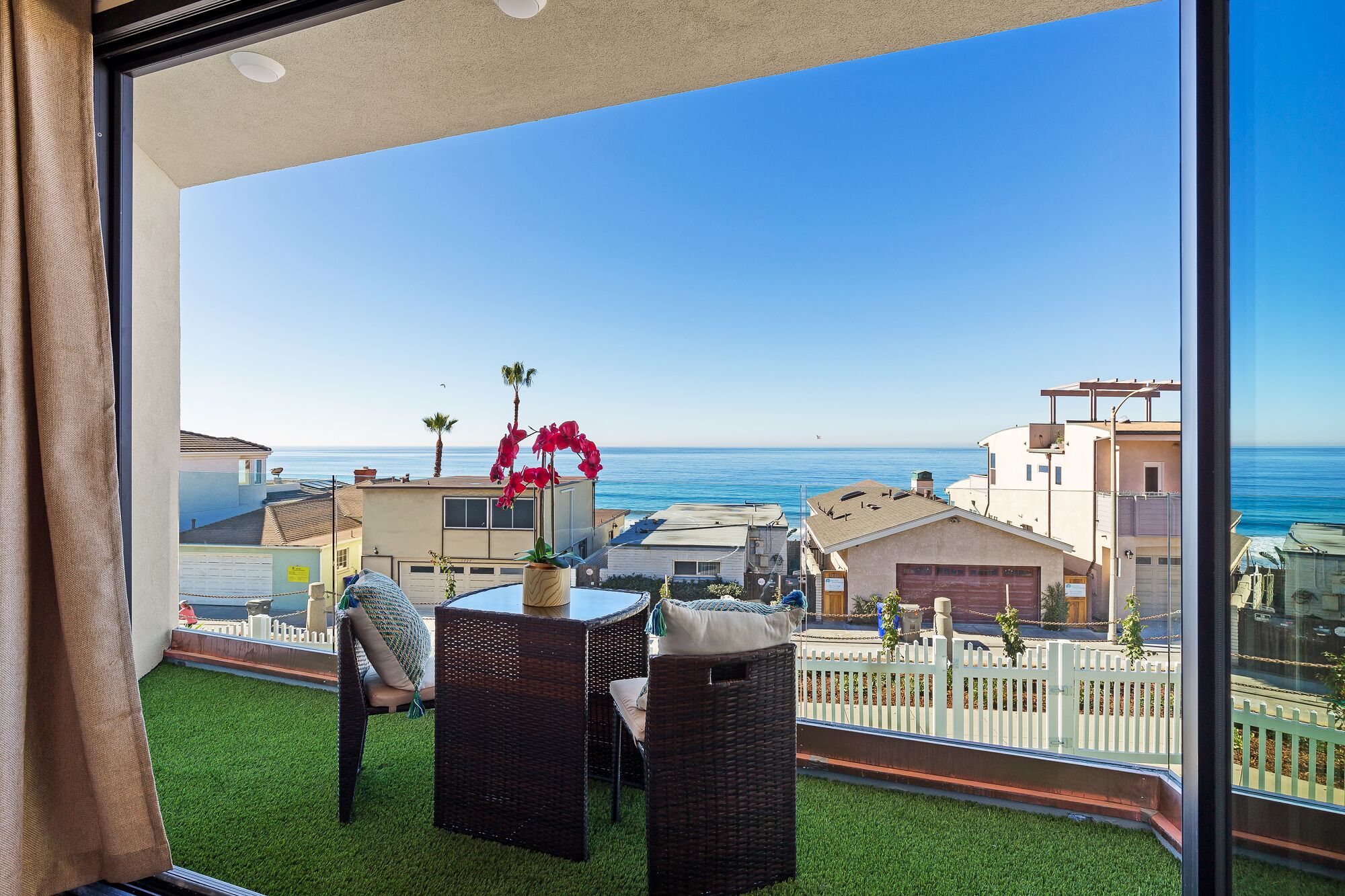 A balcony with wicker furniture, a small table with a vase of flowers, and a view of the ocean and neighboring buildings.