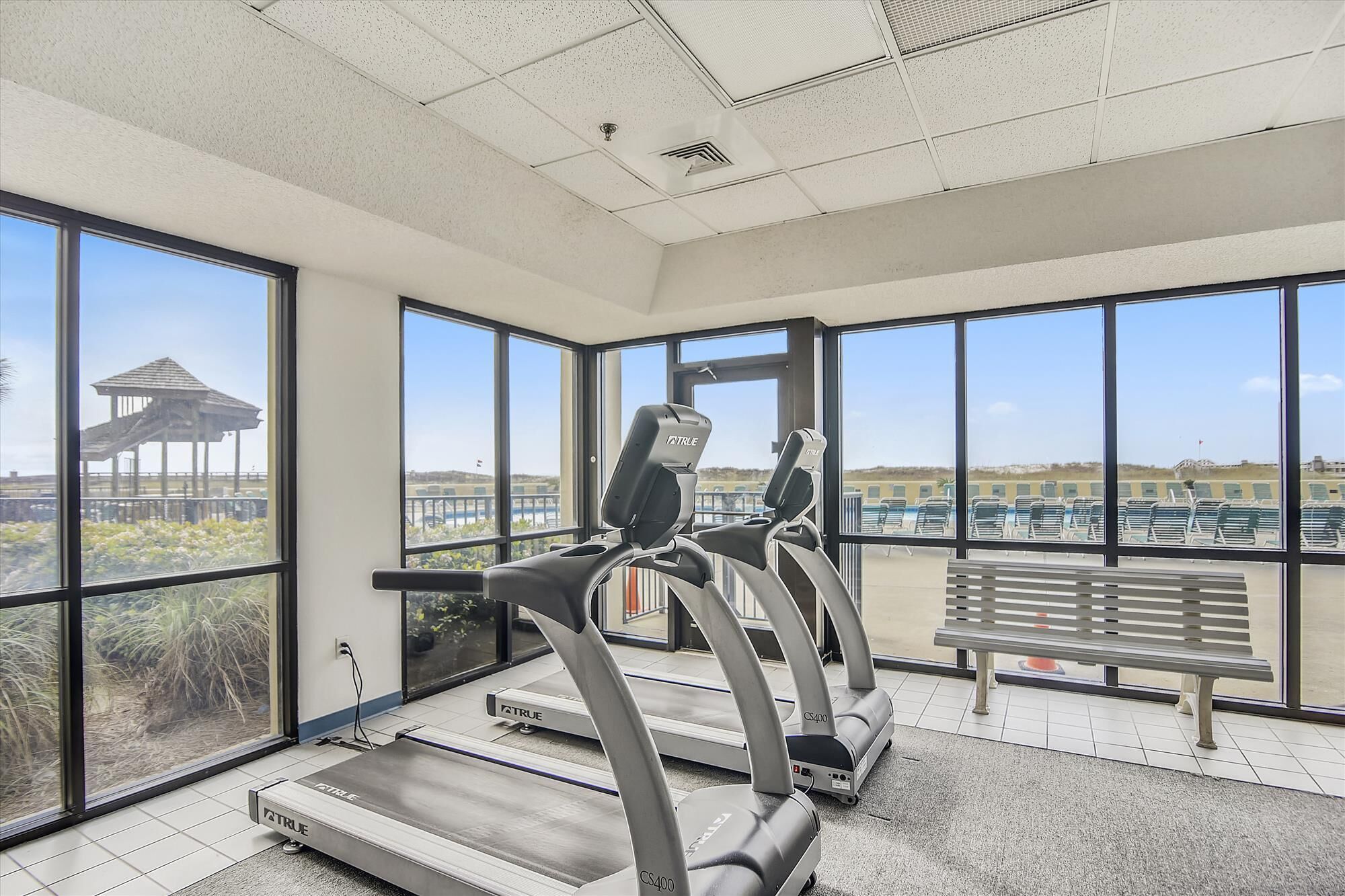 A gym with treadmills and a bench, large windows, and a view of a beach.