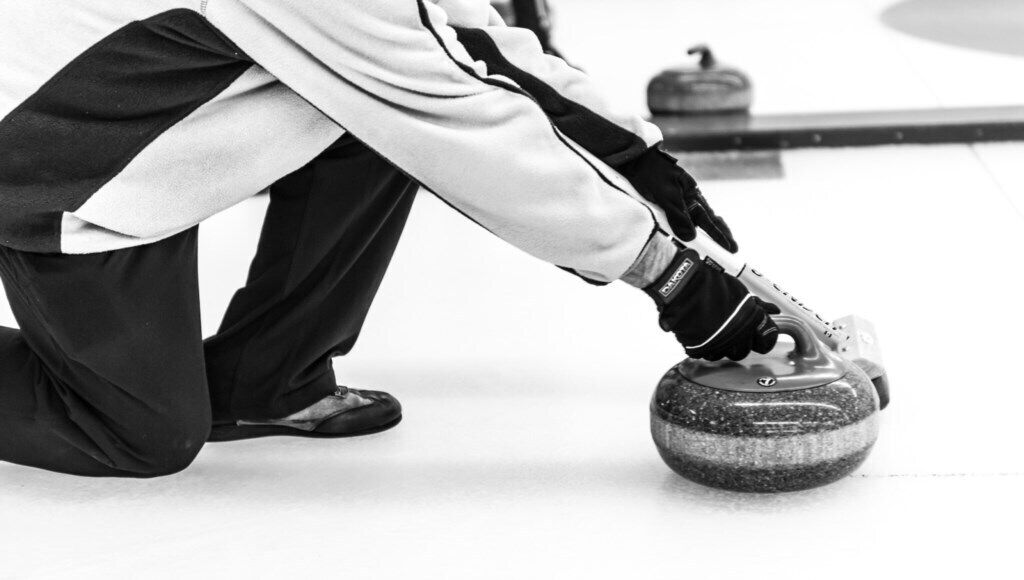 Una persona con chaqueta blanca y pantalones negros, arrodillada sosteniendo una piedra de curling.
