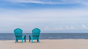 Twee turquoise Adirondack-stoelen op een zandstrand, met de oceaan op de achtergrond.