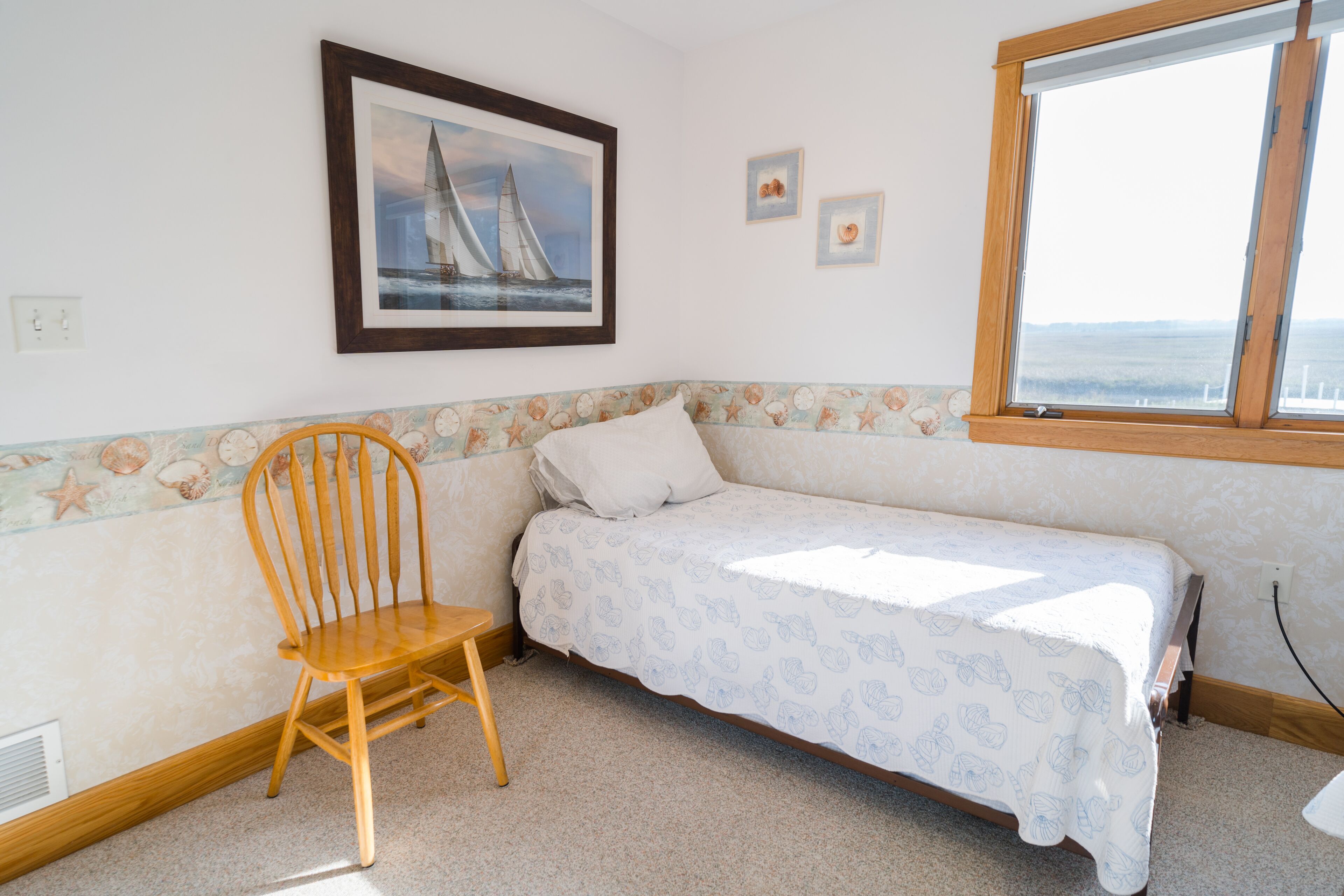 A bedroom with a single bed, a wooden chair, a framed picture of a sailboat, and a window with a view.