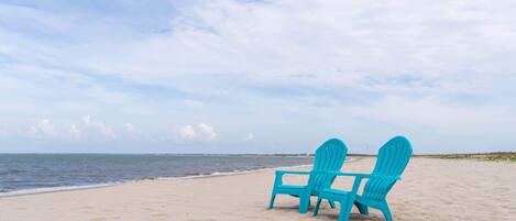 Two turquoise Adirondack chairs on a sandy beach with a view of the ocean and a cloudy sky.