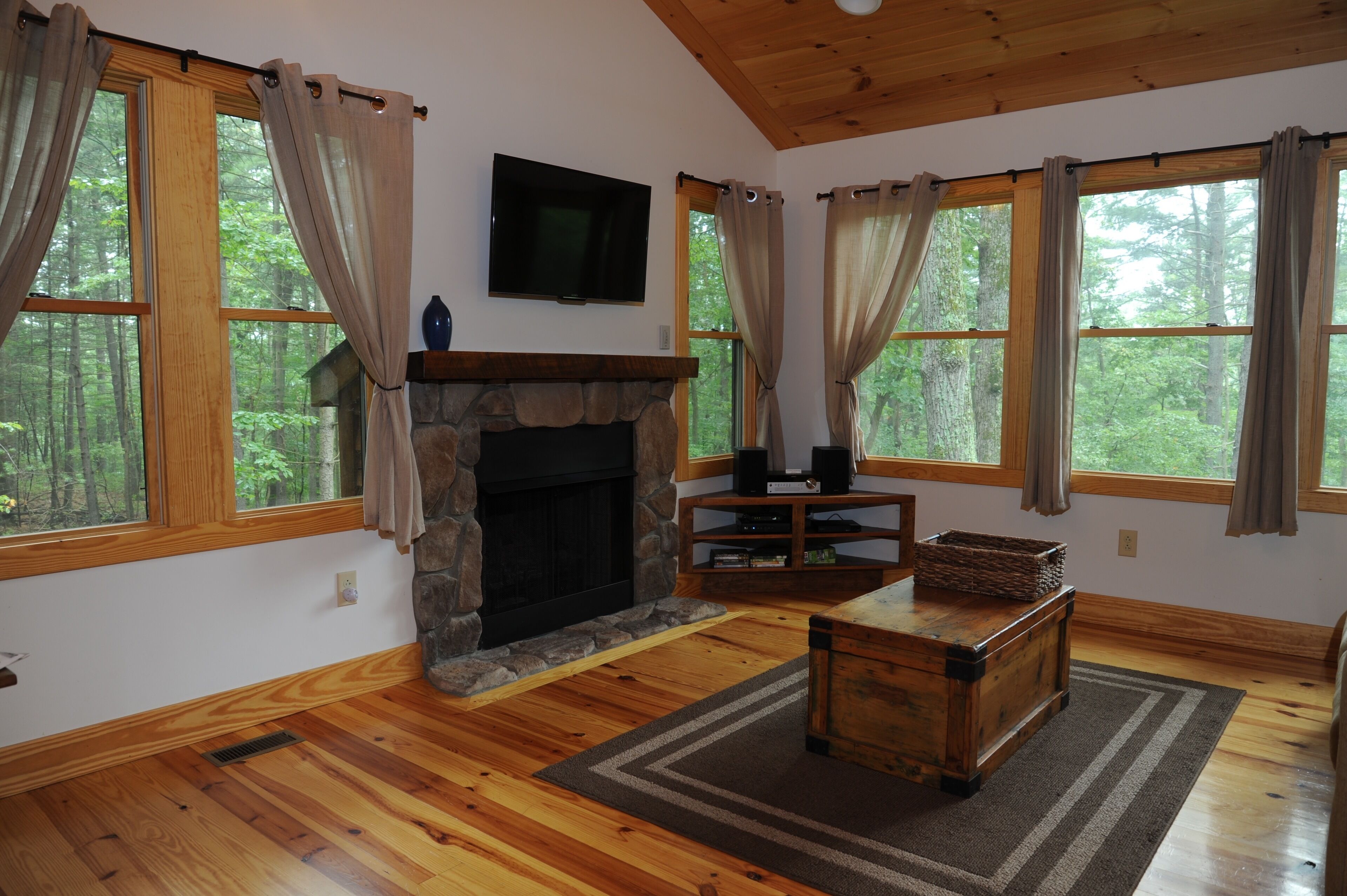 A cozy living room with a stone fireplace, wooden floors, and large windows with curtains.