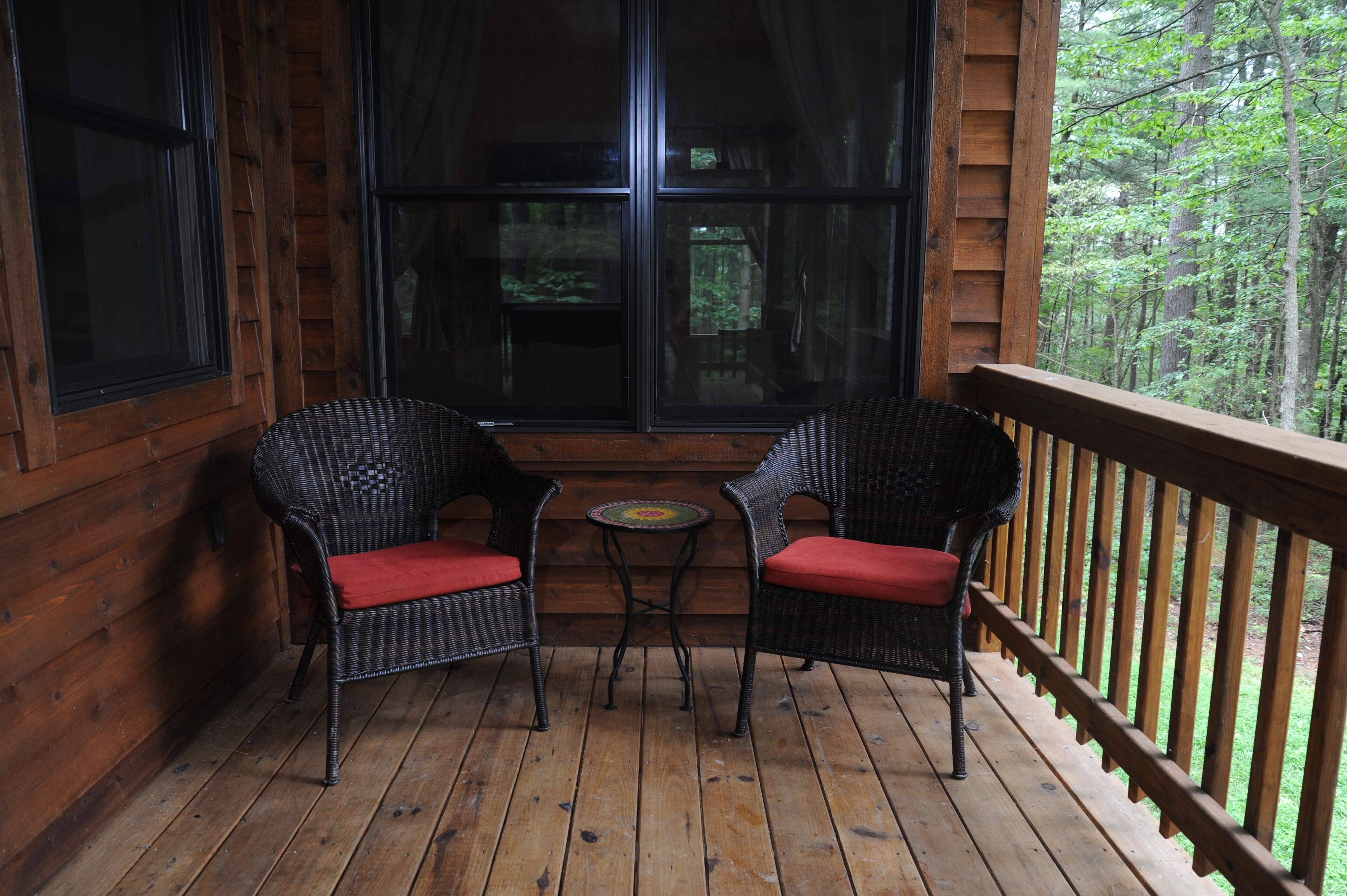Een houten terras met twee rieten stoelen en een kleine tafel, met uitzicht op een bebost gebied.