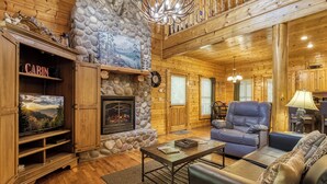 A cabin interior with a stone fireplace, wooden walls, a television, a leather chair, and a dining table. - Amazing Luxury Log Cabin! Great Living Space! Mountain Views! 2 Jacuzzi Tubs!  (Sevierville)