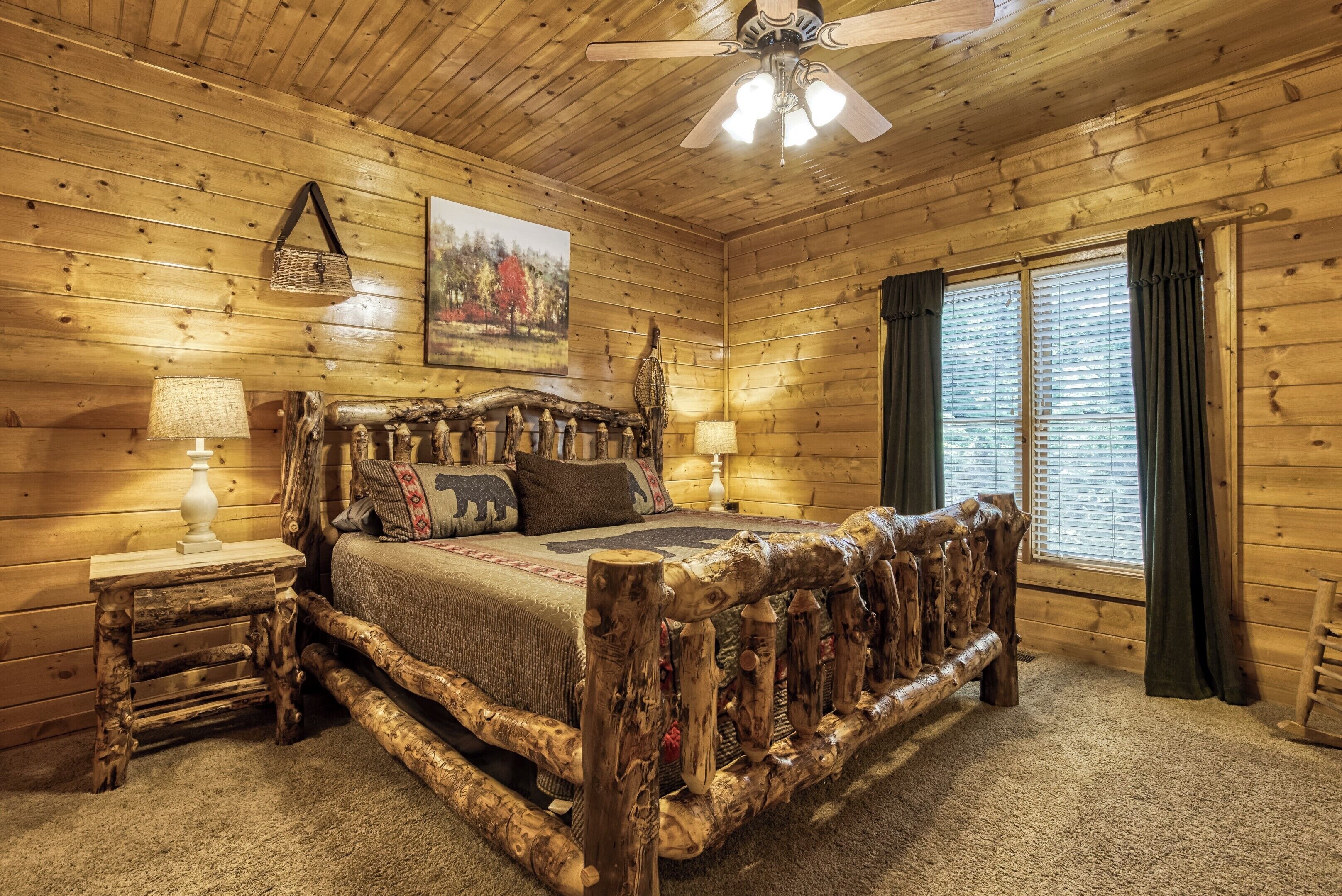 A log cabin bedroom with a rustic bed frame, wooden walls, a ceiling fan, and a window with curtains.