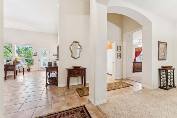 Uma sala de estar ampla com piso ladrilhado, uma mesa de centro de madeira, uma mesinha lateral e um espelho grande na parede.