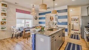 Cocina moderna con una isla central, encimeras de granito y electrodomésticos de acero inoxidable. Incluye lámparas colgantes, un salpicadero a rayas y una zona de comedor con mesa y sillas.