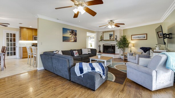 Ein Wohnzimmer mit grauem Ecksofa, einem weißen Sessel, einem Couchtisch und einem Kamin. Es gibt Deckenventilatoren, eine Küche im Hintergrund und einen Essbereich mit Stühlen.