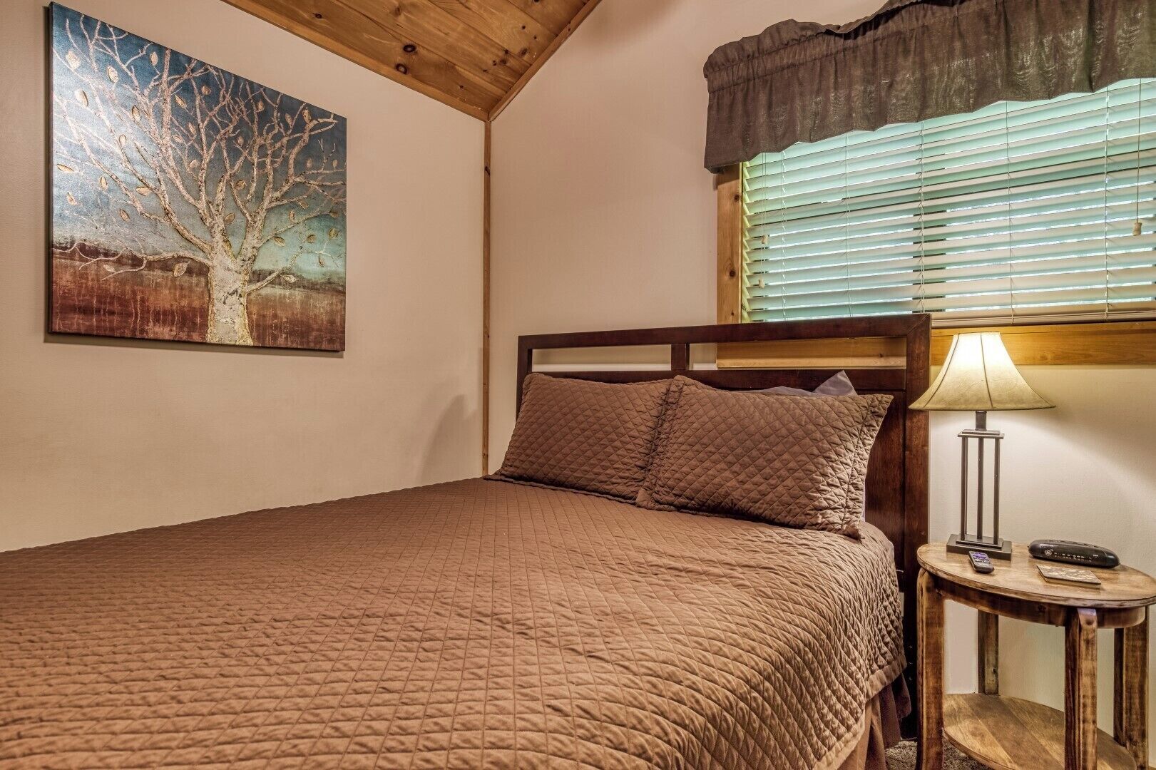 A bedroom with a wooden headboard, a quilted bedspread, a bedside lamp, and a painting of a tree on the wall.