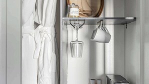 Een badkamer met een badjas aan de deur, een spiegel, een koffiezetapparaat en een plank met kopjes en een pot.