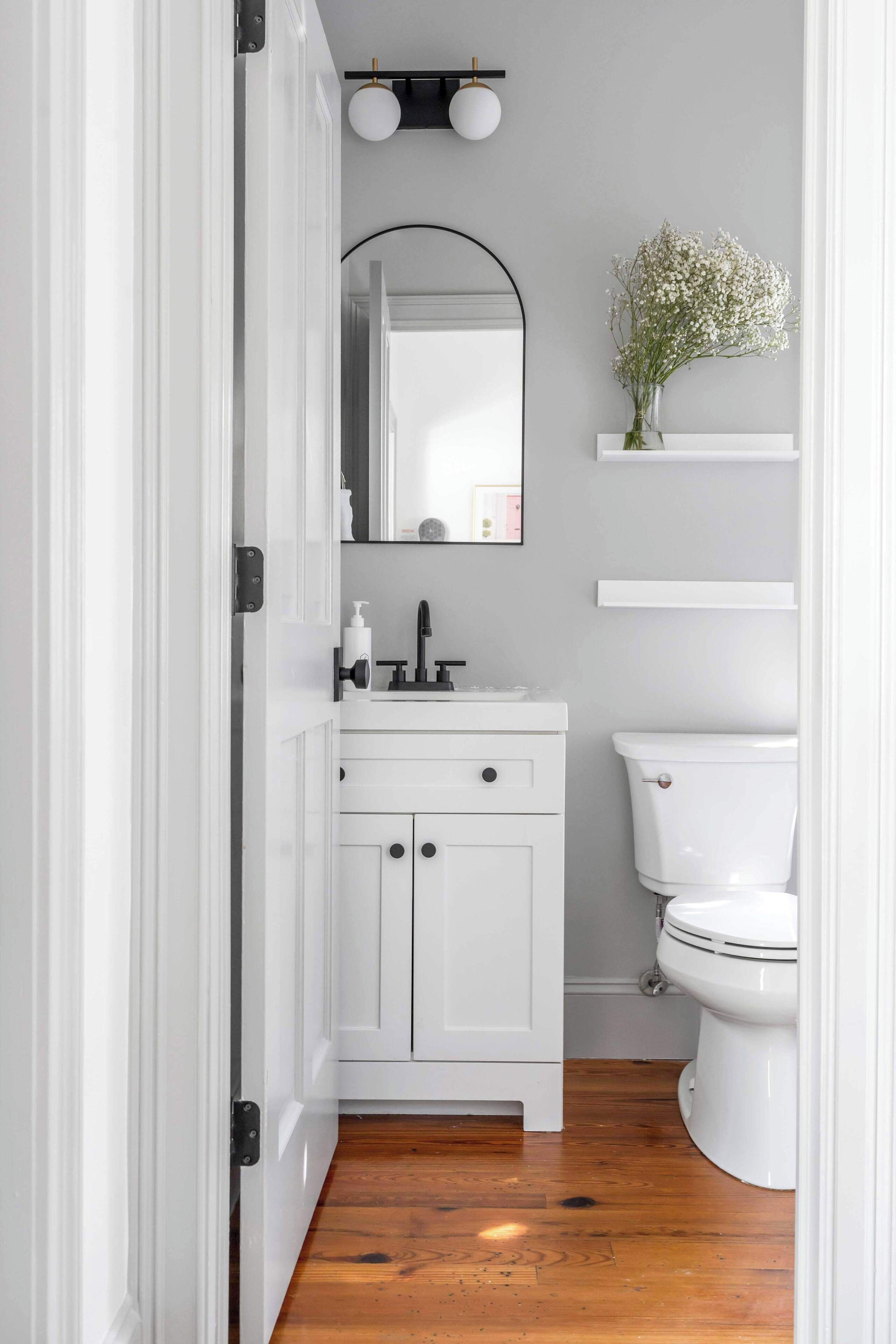 Une salle de bain avec des meubles blancs, des toilettes blanches, un miroir au-dessus du lavabo et une petite étagère sur laquelle est posé un vase de fleurs.
