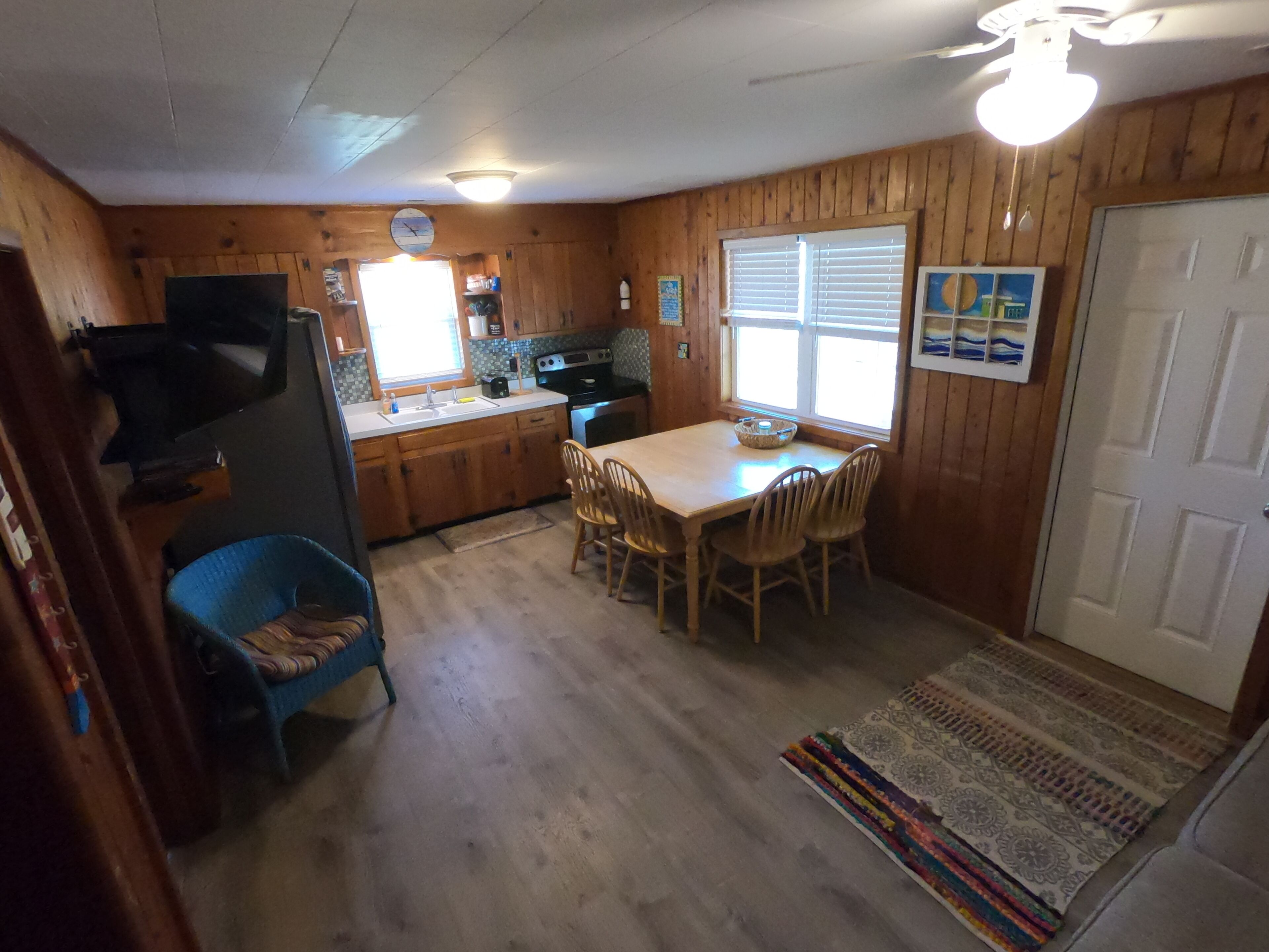 A compact kitchen with wooden cabinets, a dining table with chairs, a blue chair, a rug, and a door.