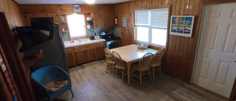 A compact kitchen with wooden cabinets, a dining table with chairs, a blue chair, a rug, and a door.