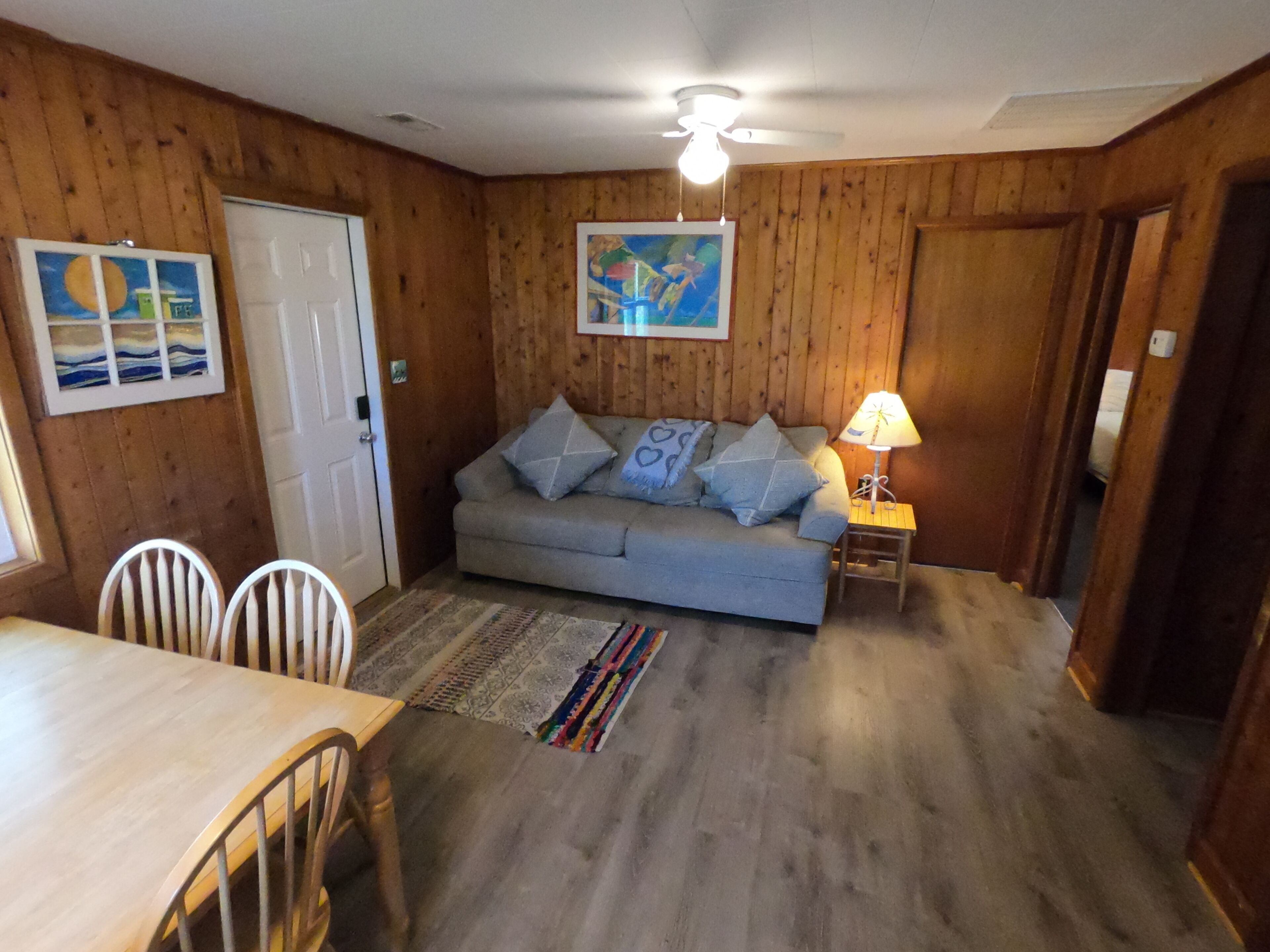 A cozy room with a wooden interior, a grey sofa with patterned cushions, a small table with a lamp, and a door.
