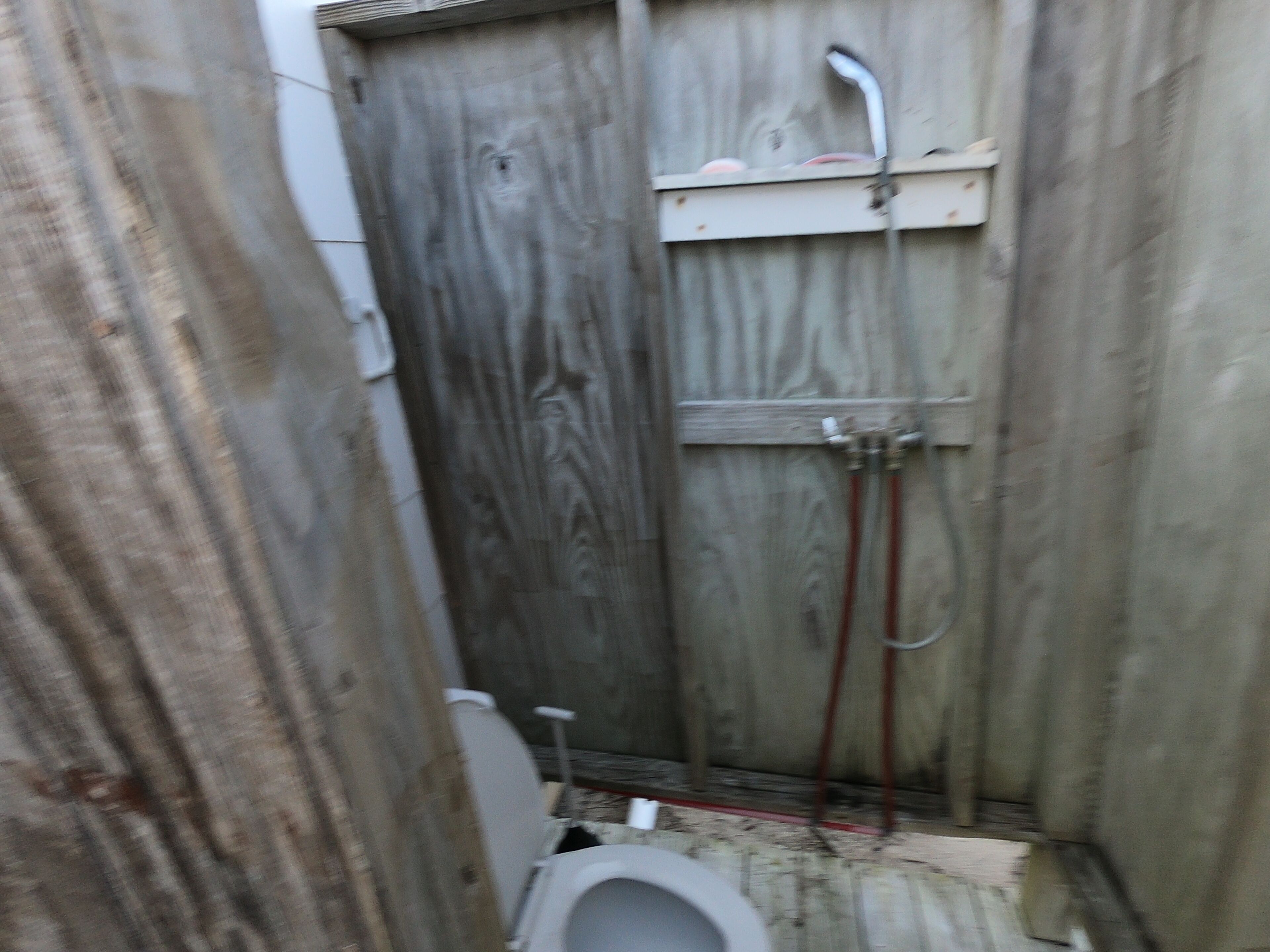 Un baño estrecho con paredes de madera, un inodoro, zona de ducha y tuberías a la vista.