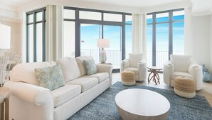 Ein geräumiges Wohnzimmer mit großem Fenster, einer Couch, zwei Sesseln, einem Couchtisch und einem kleinen Beistelltisch mit Lampe.