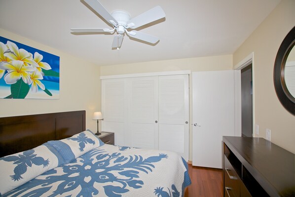 A bedroom with a bed, a ceiling fan, a painting of flowers, a wardrobe, and a dresser.