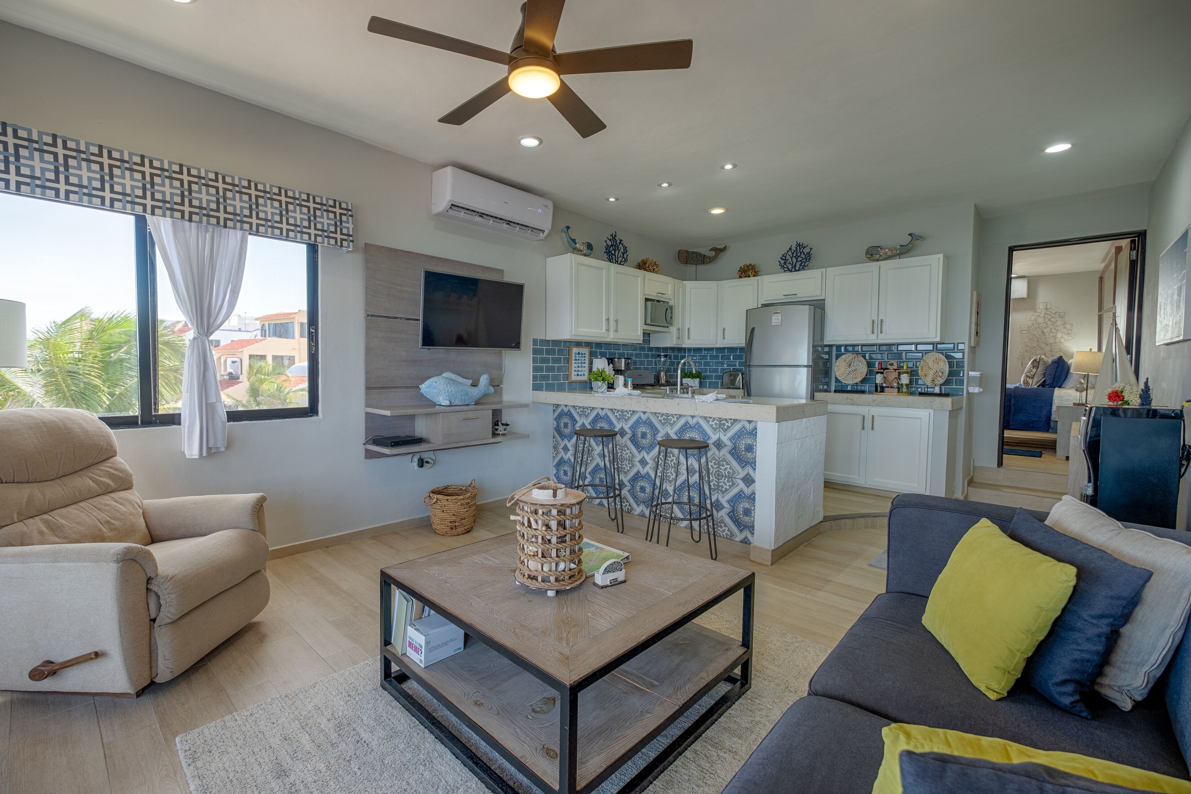 A modern living room with a sofa, a coffee table, and a TV. There is a kitchen in the background with white cabinets and a blue backsplash.