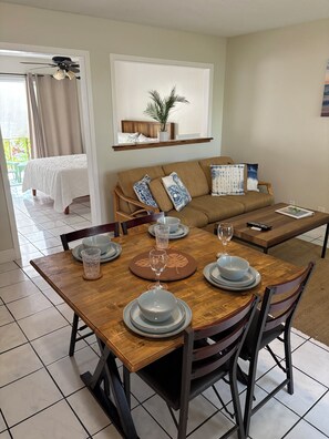 A dining area with a wooden table set for two, a sofa, and a bed in the background. - A/C and Ocean front pool, 1bedroom Condo on Ali’i drive in the heart of Kona ❤️ (Kailua-Kona)