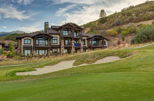 A large, multi-story house with a prominent balcony, situated on a grassy slope with a clear sky.